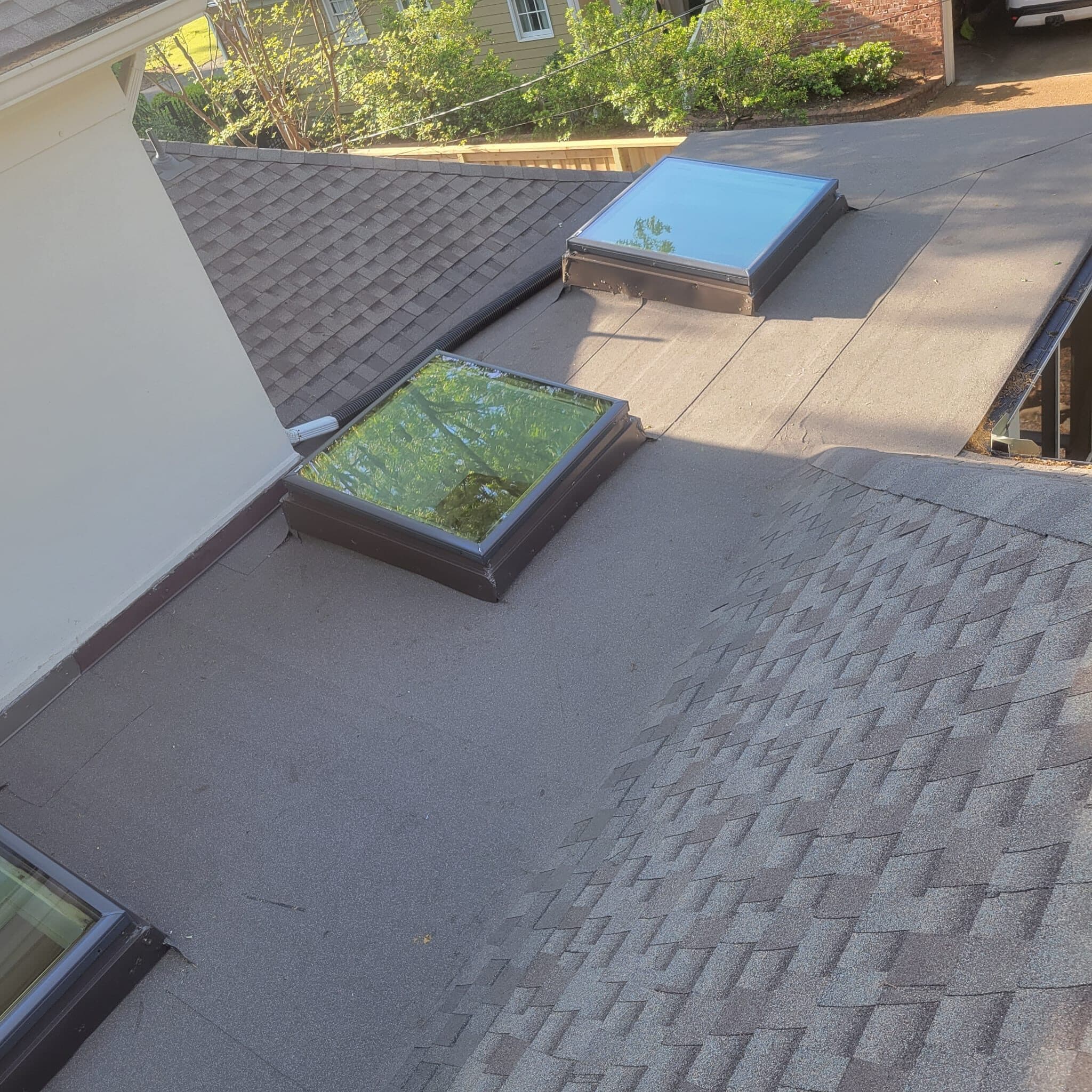 Low-slope roof section with two skylights meeting architectural shingle slopes above a wooded backyard