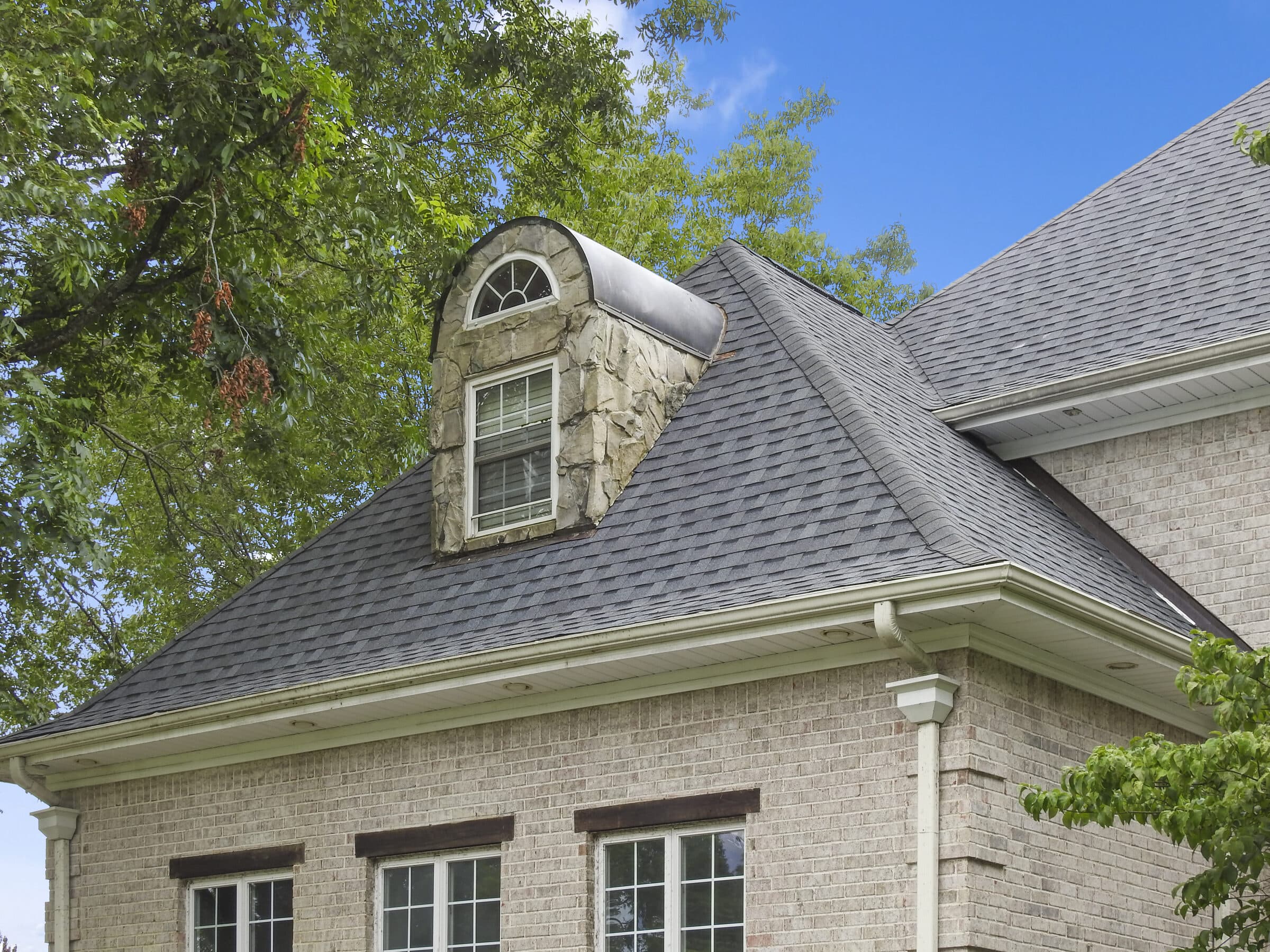Gray shingle roof and dormer on a stone home under a blue sky