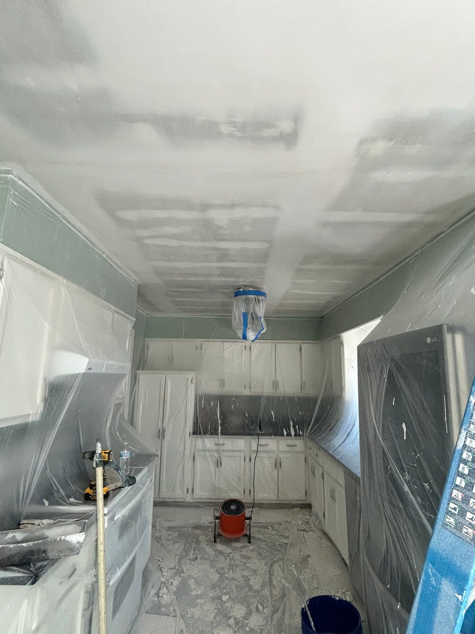 Kitchen ceiling being refinished with cabinets and counters masked in plastic and a drying fan running