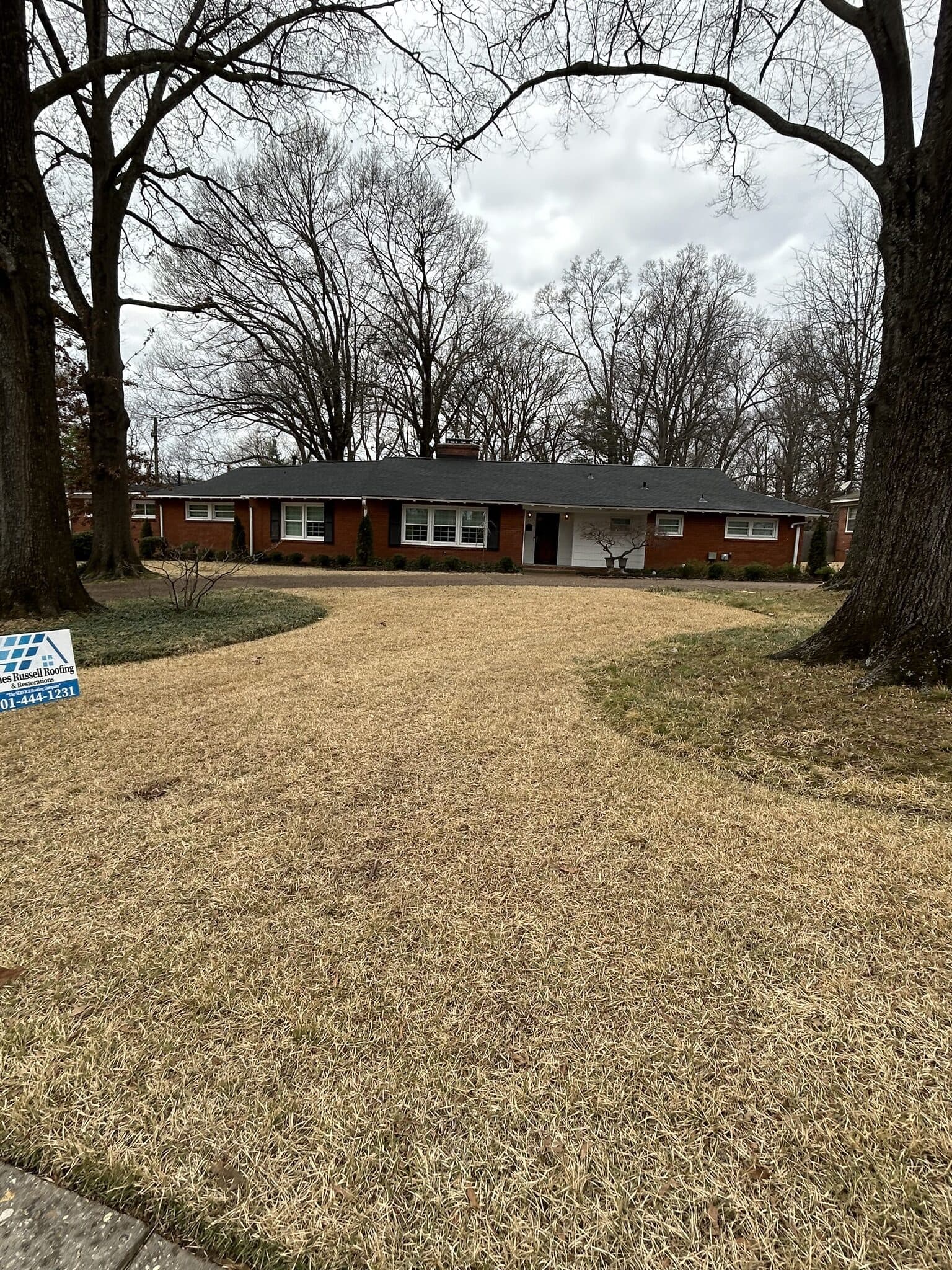 Long ranch home beneath mature trees with a company yard sign after roof completion