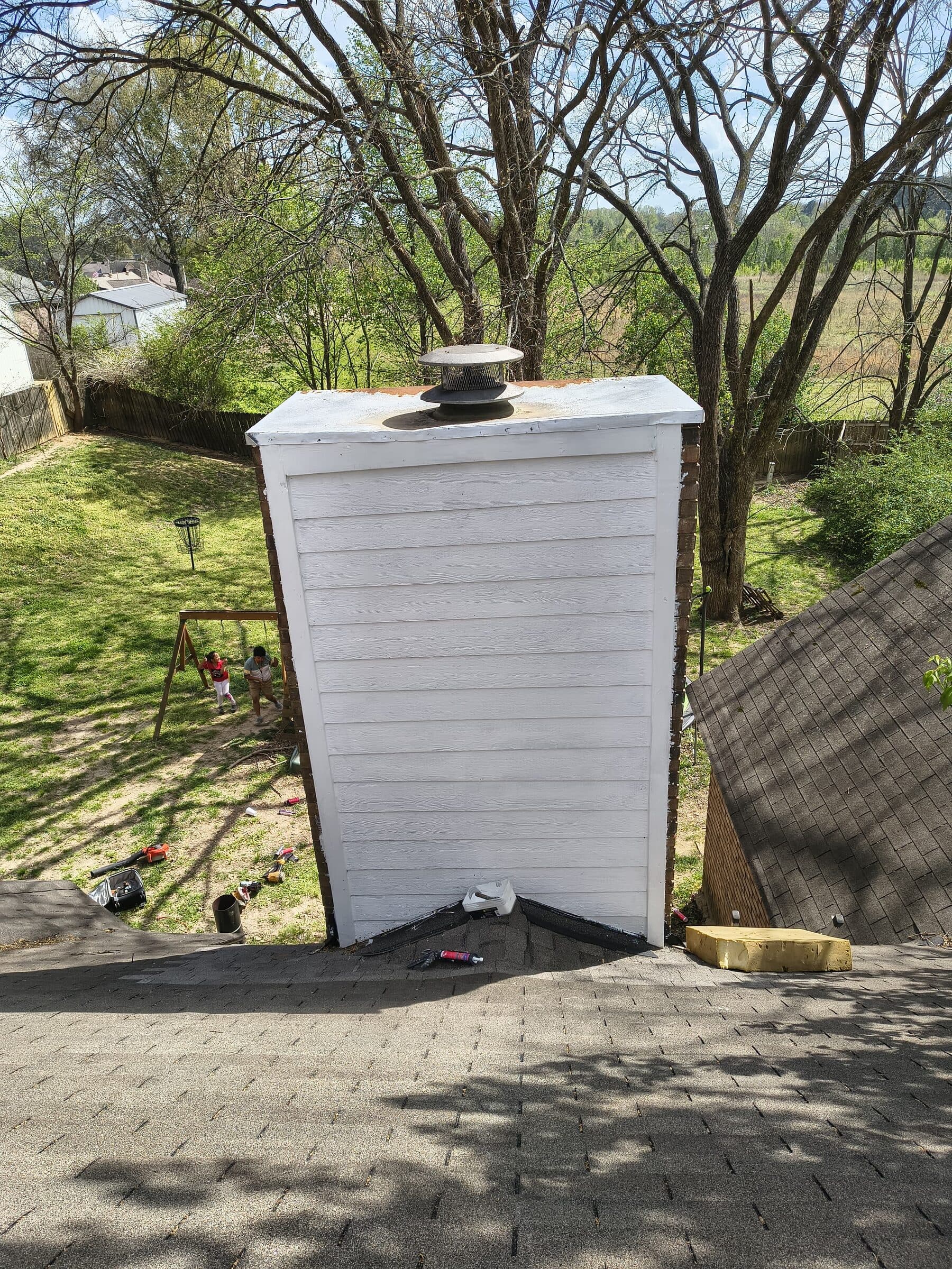 Tall white chimney with fresh flashing on a shingle roof in a wooded lot