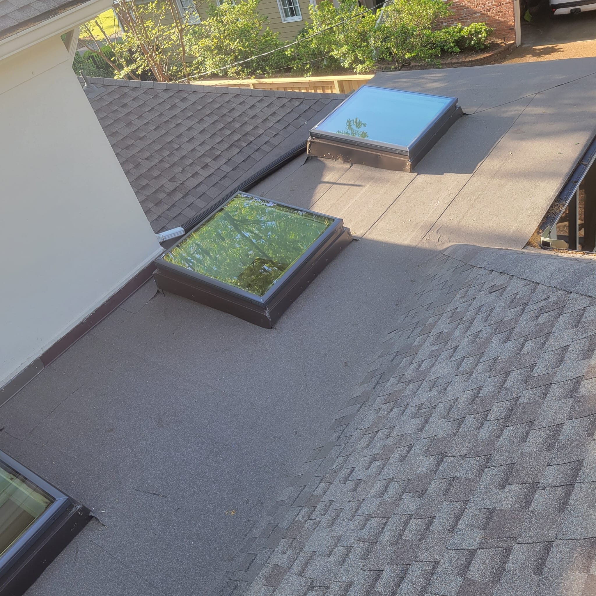Low-slope roof section with two skylights meeting architectural shingle slopes above a wooded backyard