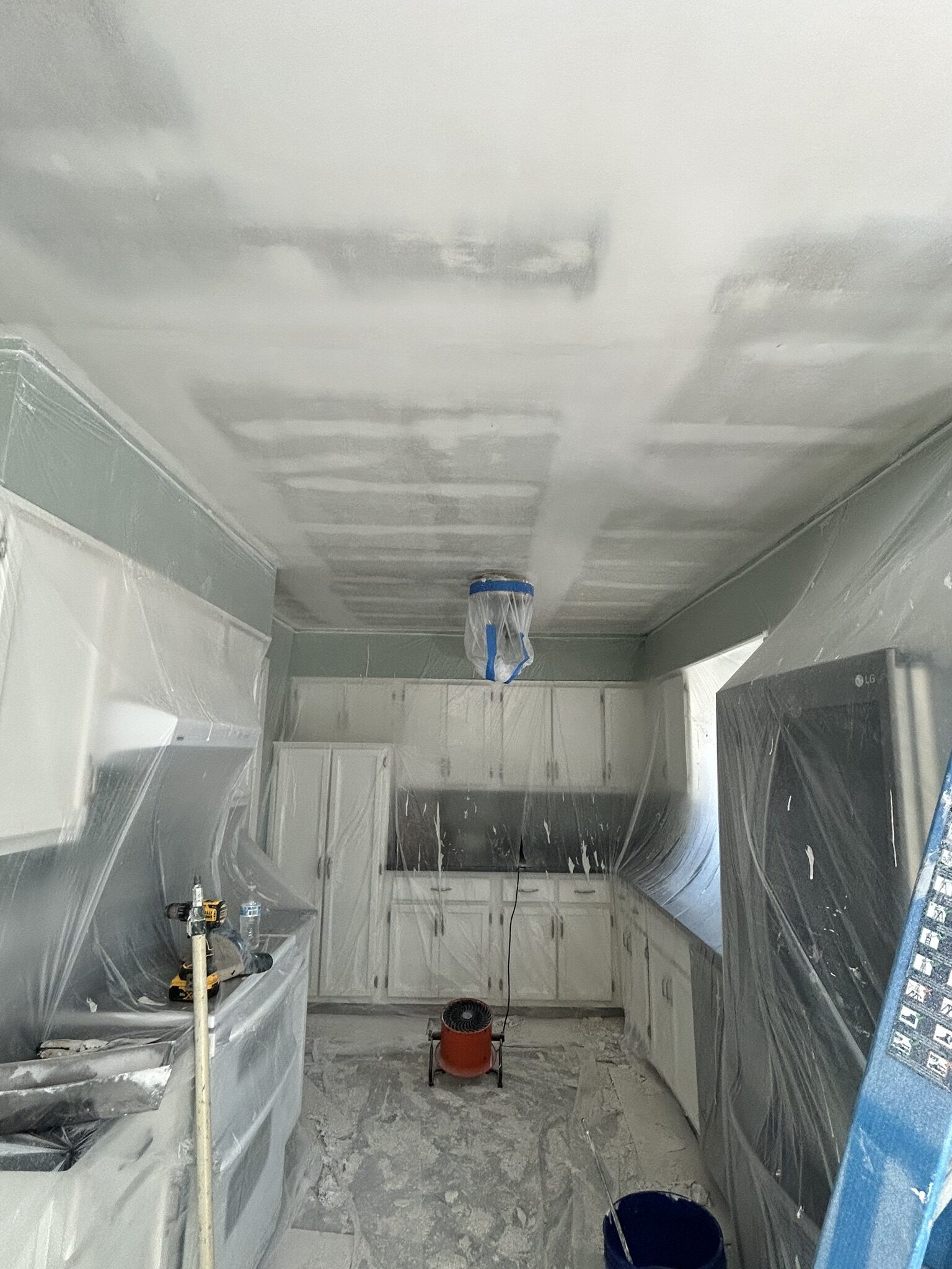 Kitchen ceiling being refinished with cabinets and counters masked in plastic and a drying fan running